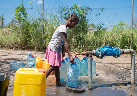 Jente fra Mosambik med fletter, hvit t-skjorte og rosa skjørt fyller vann på ei plastkanne, fra en vannpost. Foto: Cornel van Heerden