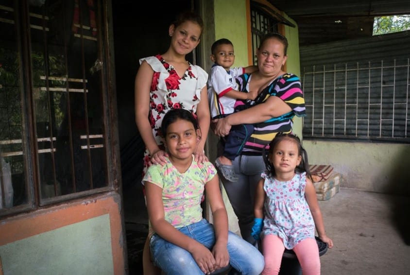 En familie i SOS-barnebyers familieprogram. En mor og tre døtre, og en sønn som hun bærer på armen, foran døra i huset de bor. Foto: Kaia Means