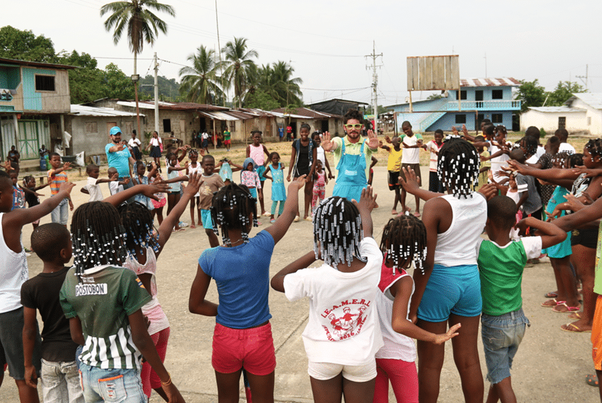 Mange barn fra Colombia står i en ring og strekker armene i været. En medarbeider fra SOS-barnebyer med blå vest med logo, leder leken. Foto SOS-barnebyer Colombia