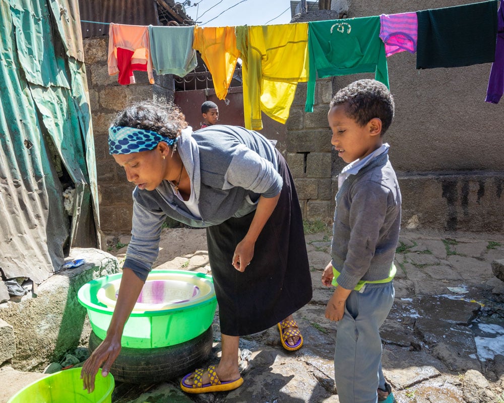 Etalem vasker klær i ei grønn balje. En av sønnene står og ser på. I bakgrunnen er det ei tørkesnor med klær. Foto: Peterik Wiggers