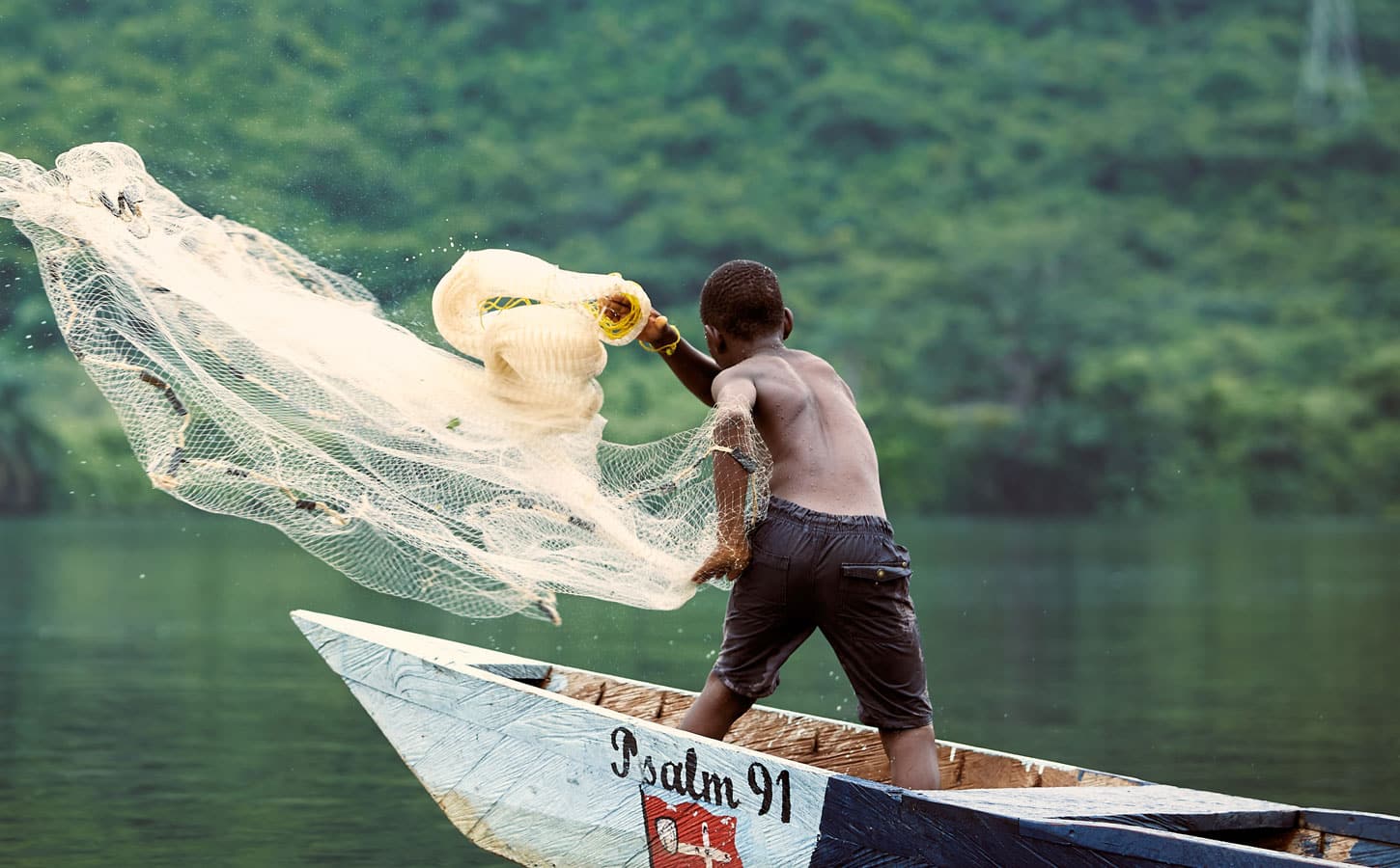 Gutt, med bar overkropp og shorts, står i en båt og kaster ut et garn. Foto: Henry KD