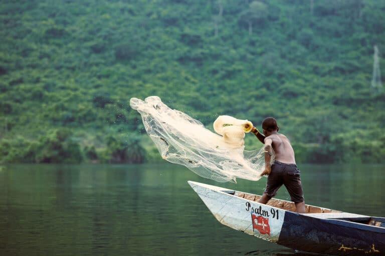 Gutt, med bar overkropp og shorts, står i en båt og kaster ut et garn. Foto: Henry KD
