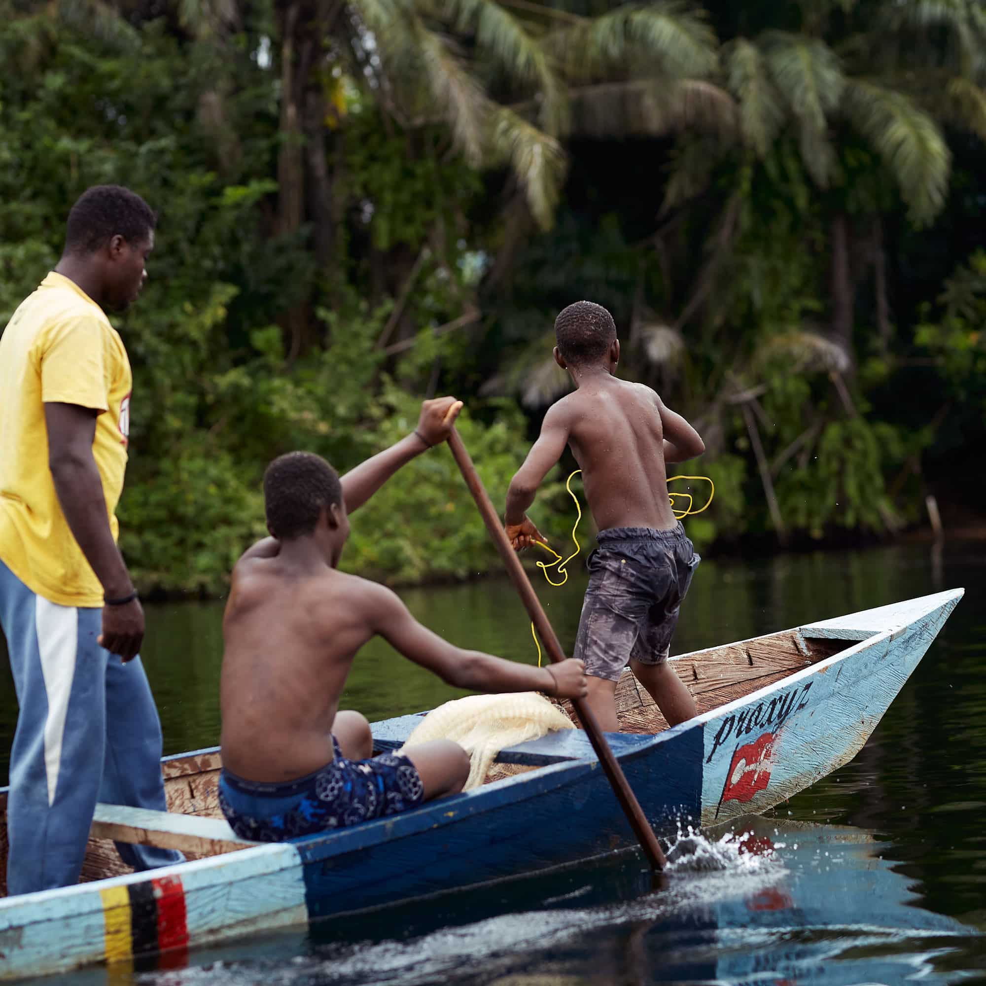 Mann i gul t-skjorte og blå joggebukser står bakerst i en båt. En gutt padler, men en annen kaster ut et fiskegarn. Foto: Henry KD