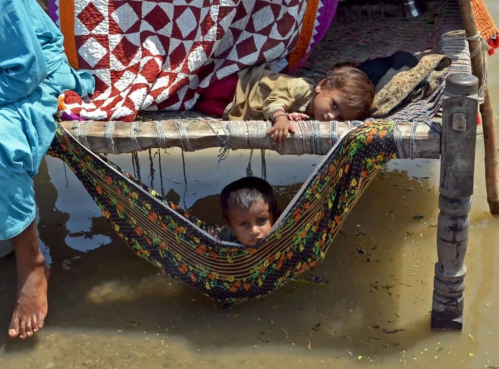 Fra flommen i Pakistan. Et lite barn ligger i en seng som står på høye ben i vannet. Det er festet et klede som en hengekøye under senga, der ligger et annet barn. Foto: Naseebullah Jamali