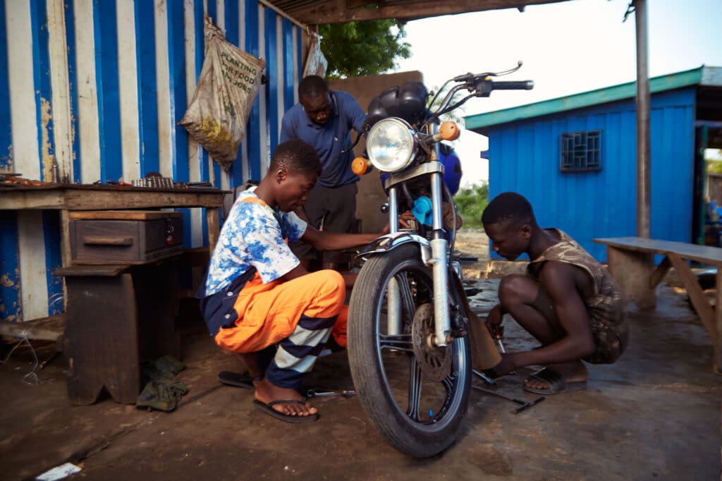 Teye sitter på huk ved siden av en motorsykkel for reparere denne. Han har på seg hvit t-skjorte og oransje selebukser. 
