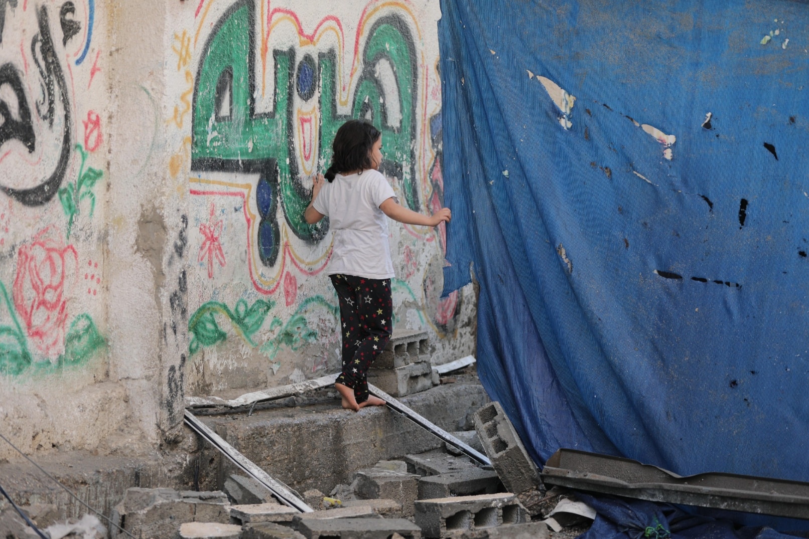 Jente med mørk bukse og hvit t-skjorte står inntil en vegg og titter bak en blå, ødelagt presenning. Foto Salah Hosny