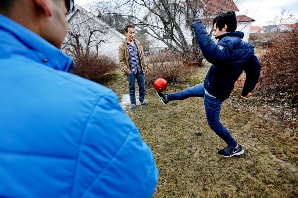 Fotballspill i hagen, med fosterfar og to av hans fostersønner. Den ene gutten i mørk blå jakke sparker en rød fotball opp i luften, mens de to andre står og ser på. Fosterfar i brun jakke, og blårutetskjorte, fosterbror står med ryggen til i lyseblå jakke.