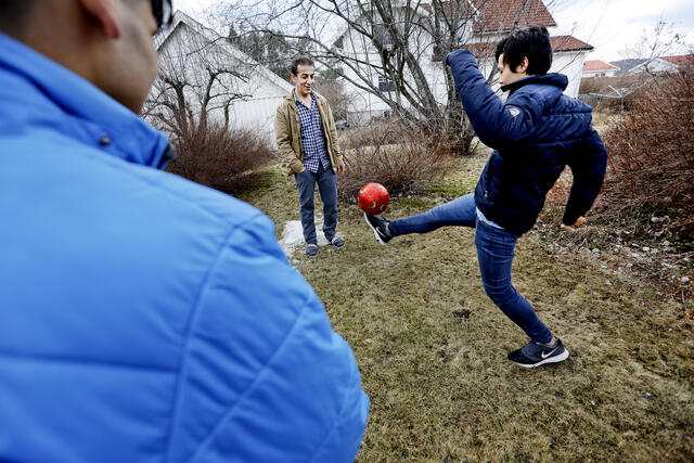 To gutter med mørkt hår og blå dunjakker sparker fotball med fosterfaren sin i hagen.