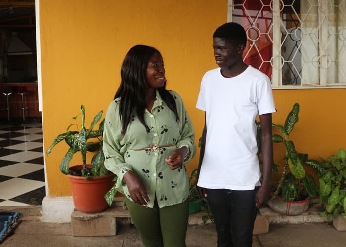 SOS-mor Anita med grønn skjorte står foran huset sitt sammen med en av gutta, han har hvit t-skjorte og jeans. Foto: SOS-barnebyer