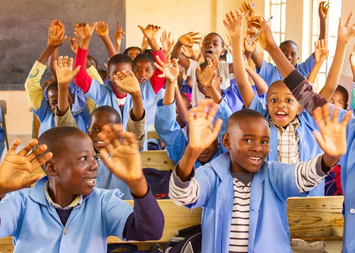 Skolegutter fra Huambo, med lyseblå skjorter som skoleuniform, sitter ved pultene og strekker hendene i været. Foto: SOS-barnebyer