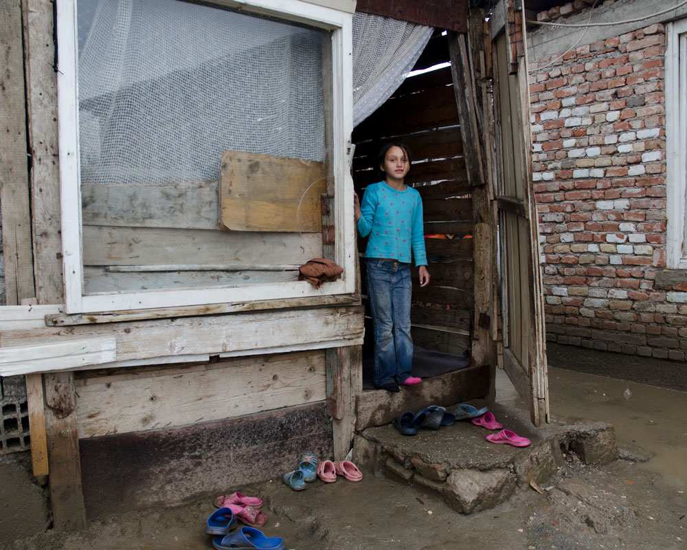 Jente med mørkt hår, turkis genser og jeans står i inngangsdøra til huset. Vegger og vinduer er i dårlig stand. Foto: Katerina Ilievska