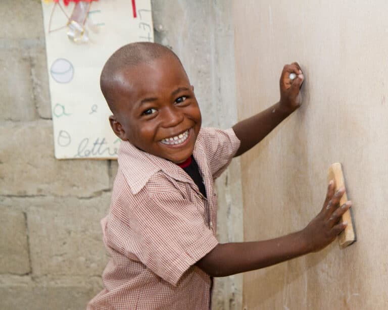 Gutt fra Haiti smårutete skjorte i rødt og hvitt pusser av ei hvit tavle. Han smiler. Foto: Daniellea Pererira