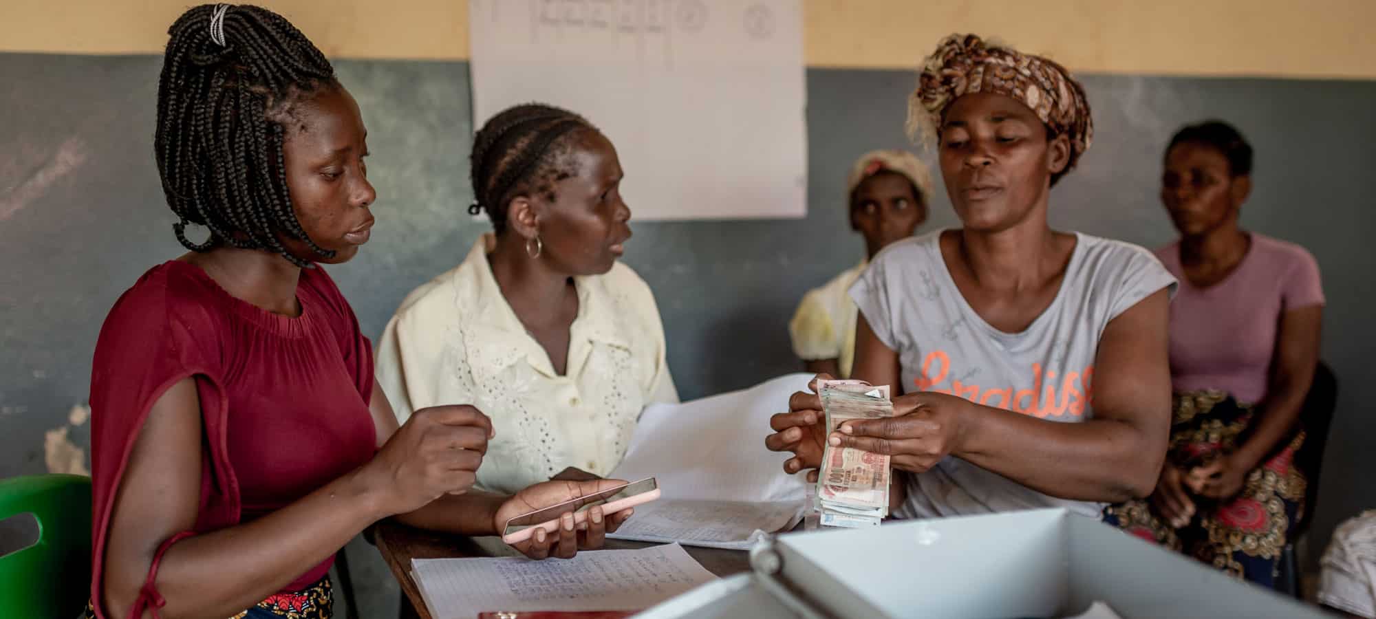Spare- og lånegrupper. Tre kvinner fra Mosambik sitter rundt et bord og teller penger, de har en kasse med lås der regnskapet og pengene oppbevares. Hun til venstre har rød kortermet genser og blomstrete skjørt, hun i midte har lys skjorte og hun til høyre har grå t-skjorte og blomstrete tørkle. Foto: Cornel van Heerden