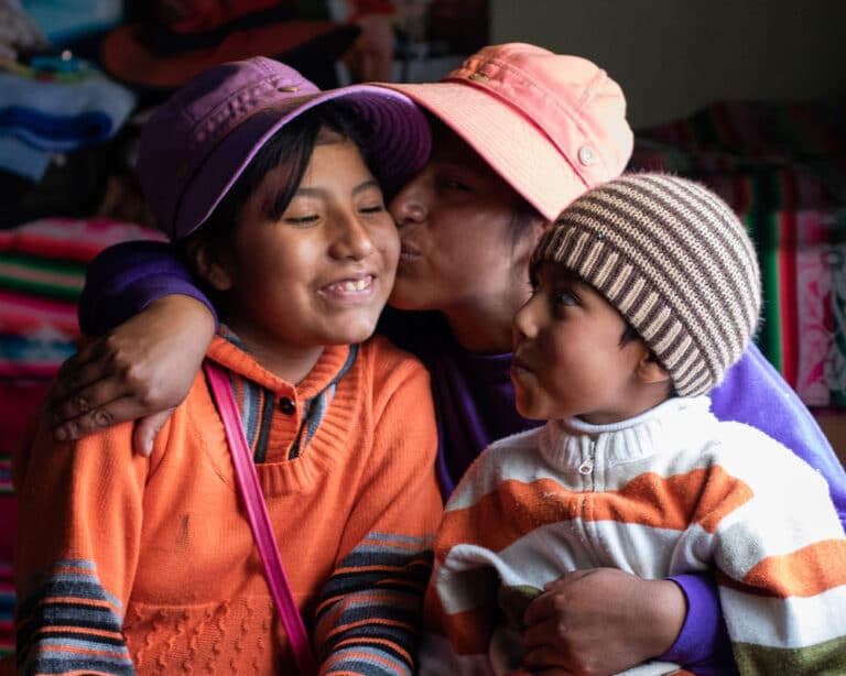 Mor fra Peru med lys oransje hatt har begge barna på fanget. Hun kysser datteren som har på seg lilla hatt og oransje genser, sønnen titter opp på dem han har stripete strikkelue og oransje og hvit stripet genser. Foto: Alejandra Kaiser