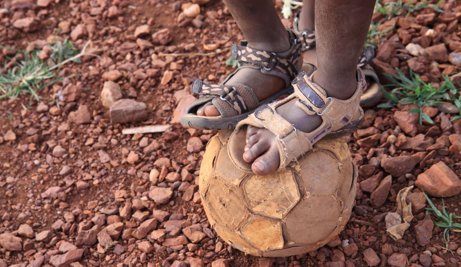 To føtter med slitte sandaler er plassert oppå en slitt fotball. Foto: Line Grove Hermansen