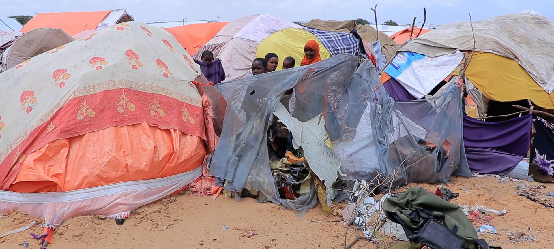 Telt i en flyktningleir dekket av tepper. Flere barn står innimellom teltene. Foto: Sunshine Media