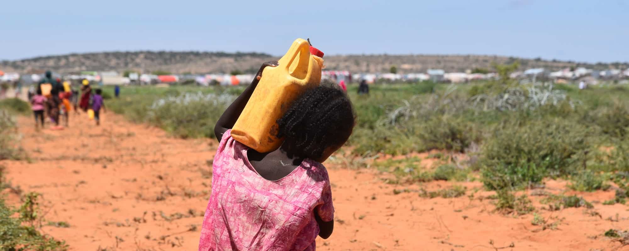 Jente fra Somaliland, med rosa kjole og flettet hår, bærer ei gul vannkanne på skuldrene. Foran henne er det en gruppe mennesker som også bærer vann og en teltleir. Foto: Carl Gustav Lorentsen