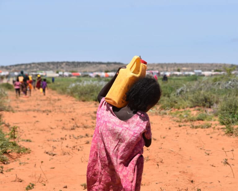 Jente fra Somaliland, med rosa kjole og flettet hår, bærer ei gul vannkanne på skuldrene. Foran henne er det en gruppe mennesker som også bærer vann og en teltleir. Foto: Carl Gustav Lorentsen