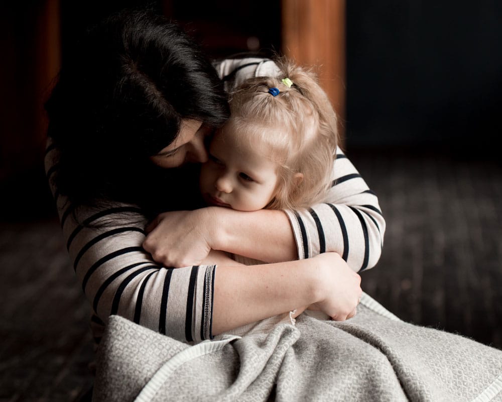 Mor fra Ukraina som har flyktet til Romania med mørkt langt hår og stripete genser holder datteren hardt inntil seg, hun er inntullet i et pledd. Foto: Katerina Ptakha
