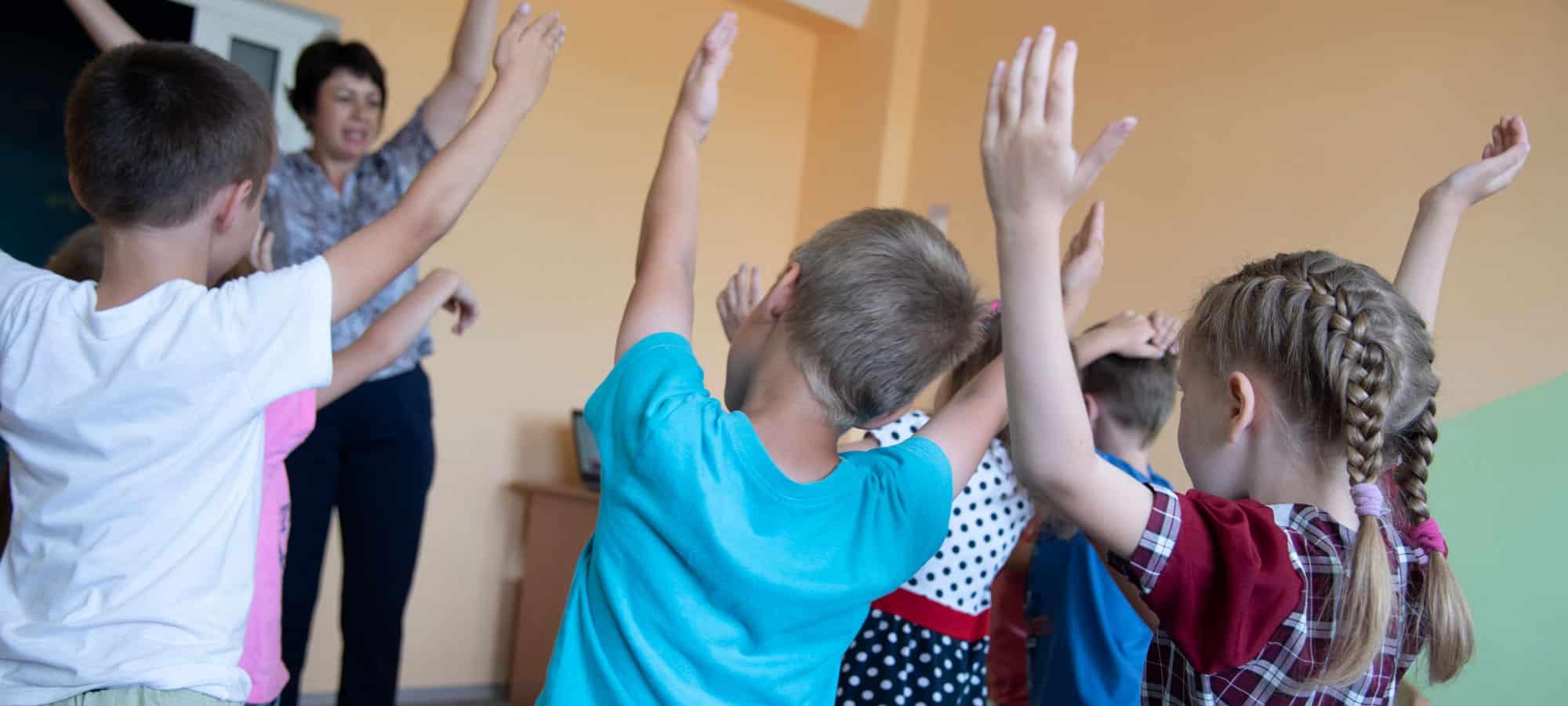 En gruppe med barn, med ryggen til, strekker hendene i været. Foran dem står psykologen, hun har mørkt kort hår, mønstret skjorte og svarte bukser. Foto: Katerina Ilievska