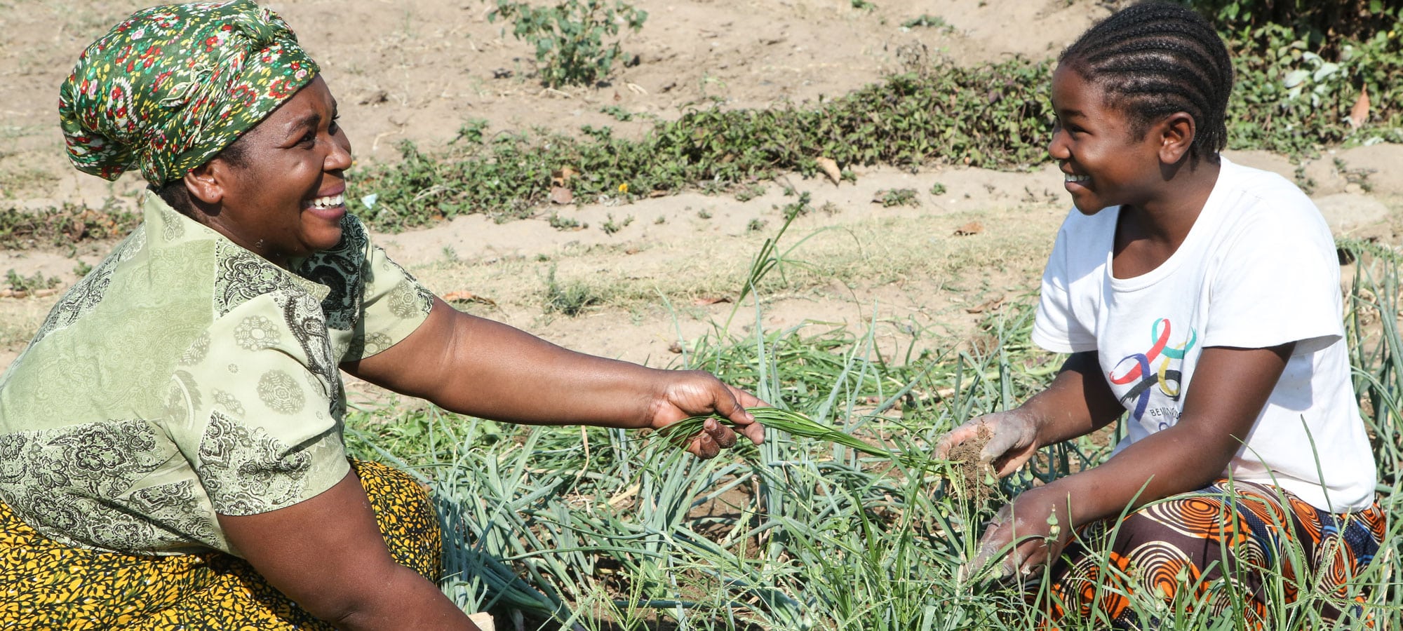 Zambia: En kvinne med mønstrete tørkle på hodet, mønstret grønn bluse og gul og svart skjørt setter planter vekster sammen med datteren med flettet hår, hvit t-skjorte og brukt møstret skjørt. Begge sitter på huk og smiler. Foto: Tom Maruko