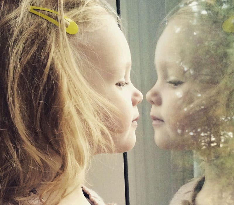 Ei lita jente med lang, lyst hår og gul spenne ser speilbildet av seg selv i et vindu. Foto: iStock