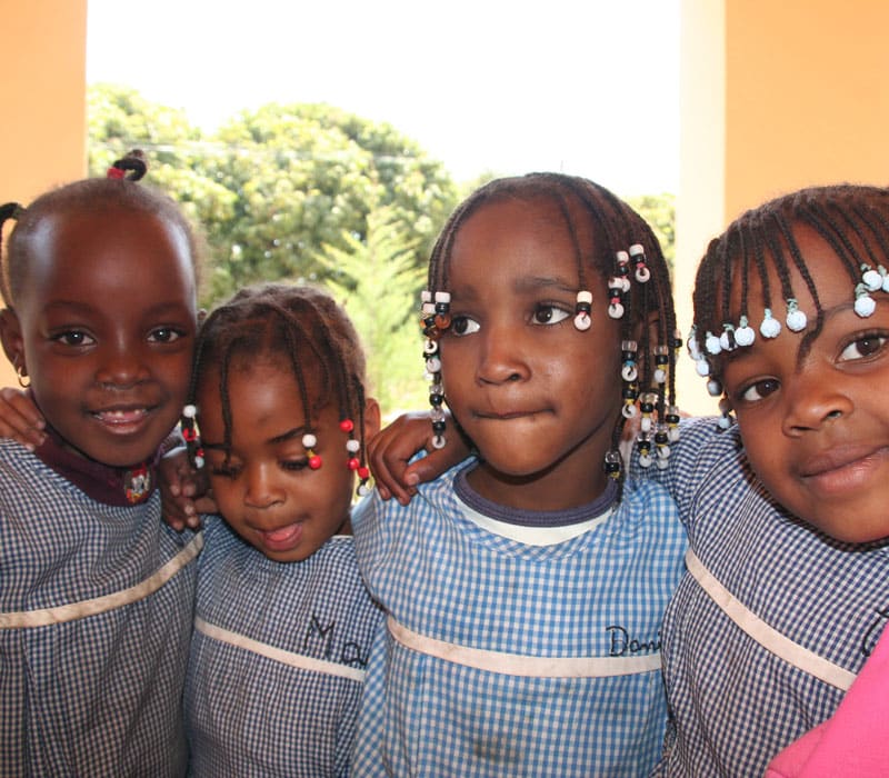 Fire jenter fra Angola har smårutete kjole, skoleuniform og fletter med perler i håret. De holder rundt hverandre. Foto: Brenda Dimbleby
