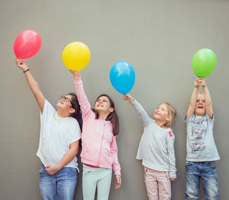 En gutt og tre jenter står inntil en grå vegg og løfter en farget ballong hver til værs. Foto: Gerhard Berger