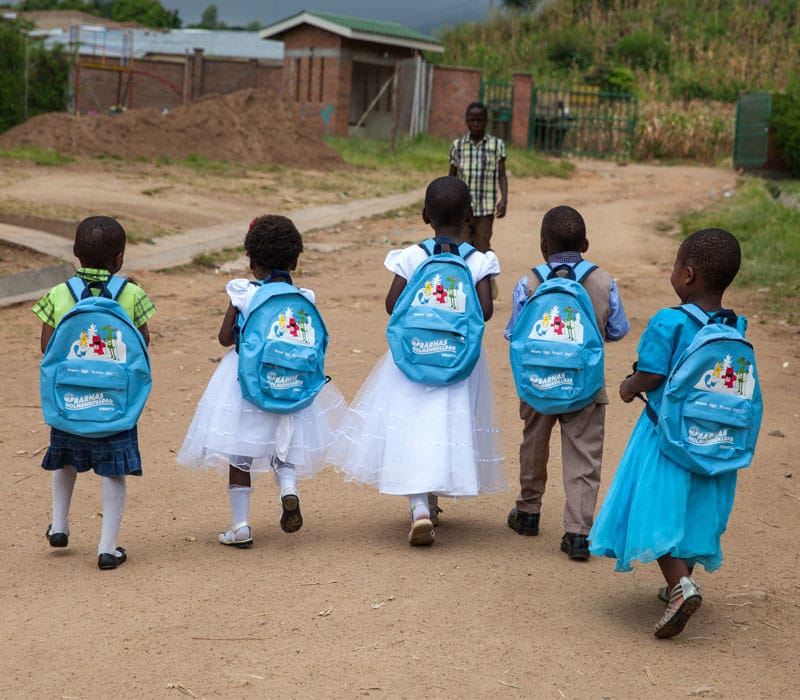 Malawi: Fire jenter og én gutt er på vei til skolen. Ei jente har grønn bluse og svart skjørt, to har hvite kjoler og ei blå. Gutten har blå skjorte og beige bukser. De går på en vei og bildet er tatt bakfra. Foto: Bjørn Owe Holmberg