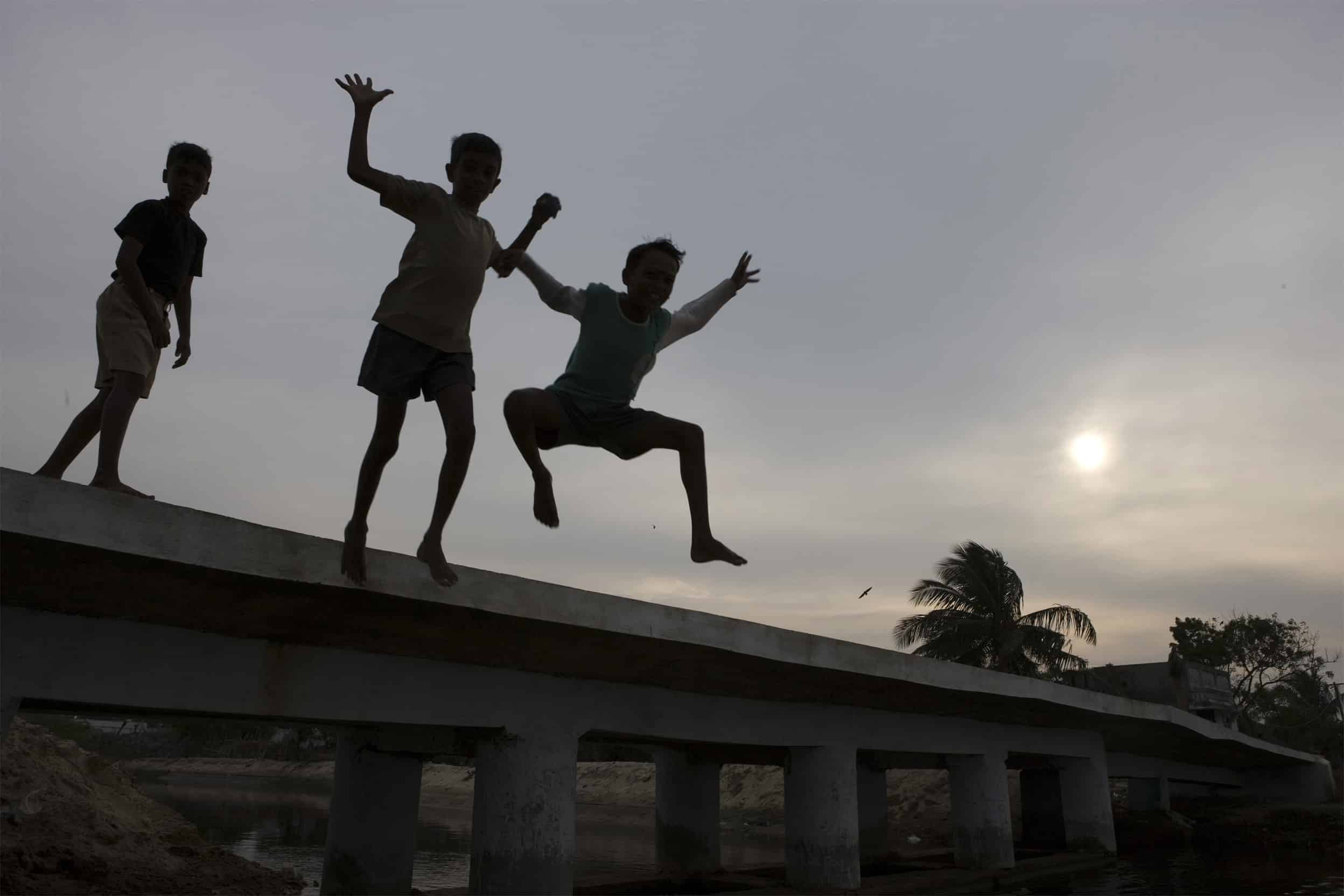 Bilde av 3 barn som hopper. De er ute og man ser solnedgang i bakgrunn.