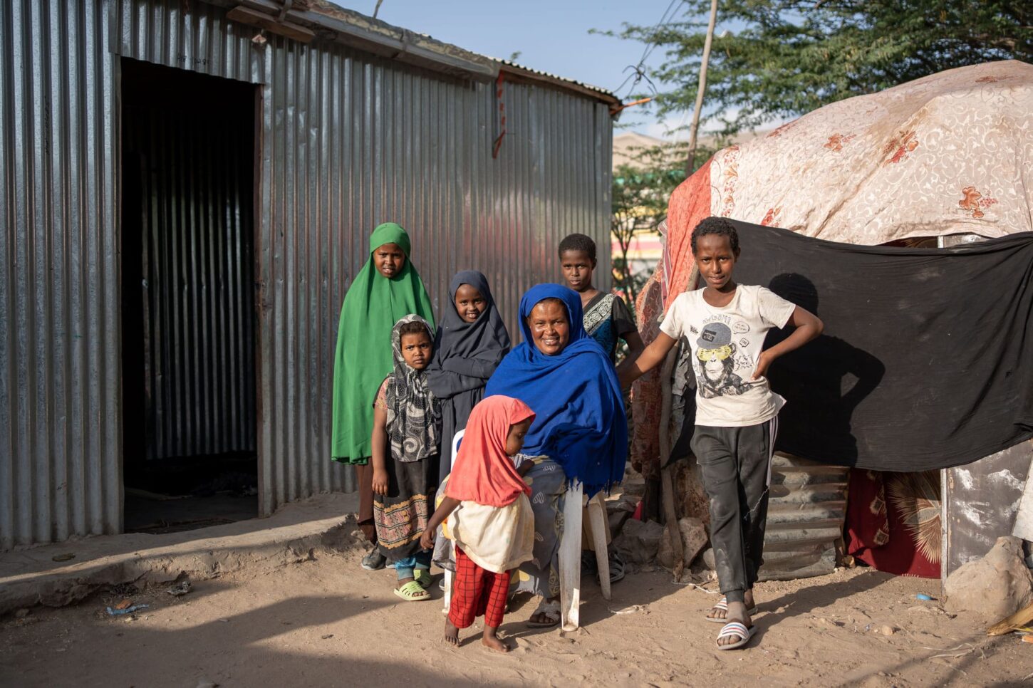 Qadan sitter utenfor det nye huset som er laget av bølgeblikkplater, de seks barna står rundt henne. Den gamle hytta, dekket av tepper, er til høyre i bildet. Foto: Lydia Mantler