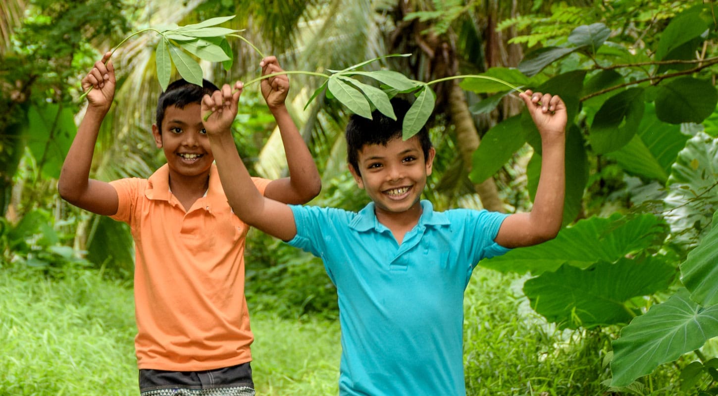 To brødre fra Sri Lanka, den ene med oransje kortermet skjorte og den andre i turkis, holder to palmeblader over hodet og smiler. Foto: Pearl Sandhu