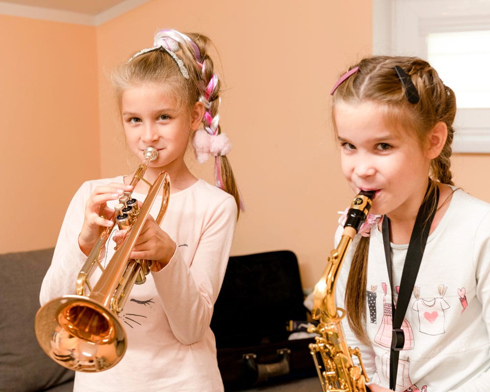 Den ene jenta, med lyst hår i ei flette med rosa bånd og rosa genser spiller trompet. Den andre med to fletter og spenner og hvit genser spiller saksofon. Foto: SOS-barnebyer