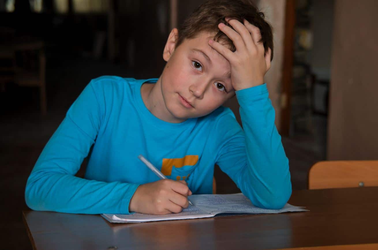 En gutt med mørkt kort hår og lys blå genser sitter ved et bord og skriver. Han støtter hodet i hånda. Foto: SOS-barnebyer