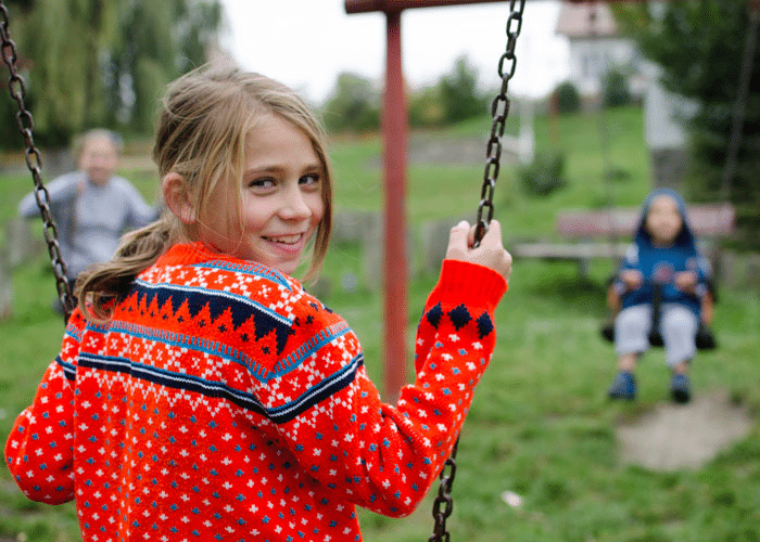 Jente med blondt, langt hår i hestehale, rød mønstret strikkejakke, sitter på ei huske og smiler. Foto: Katerina Ilievska