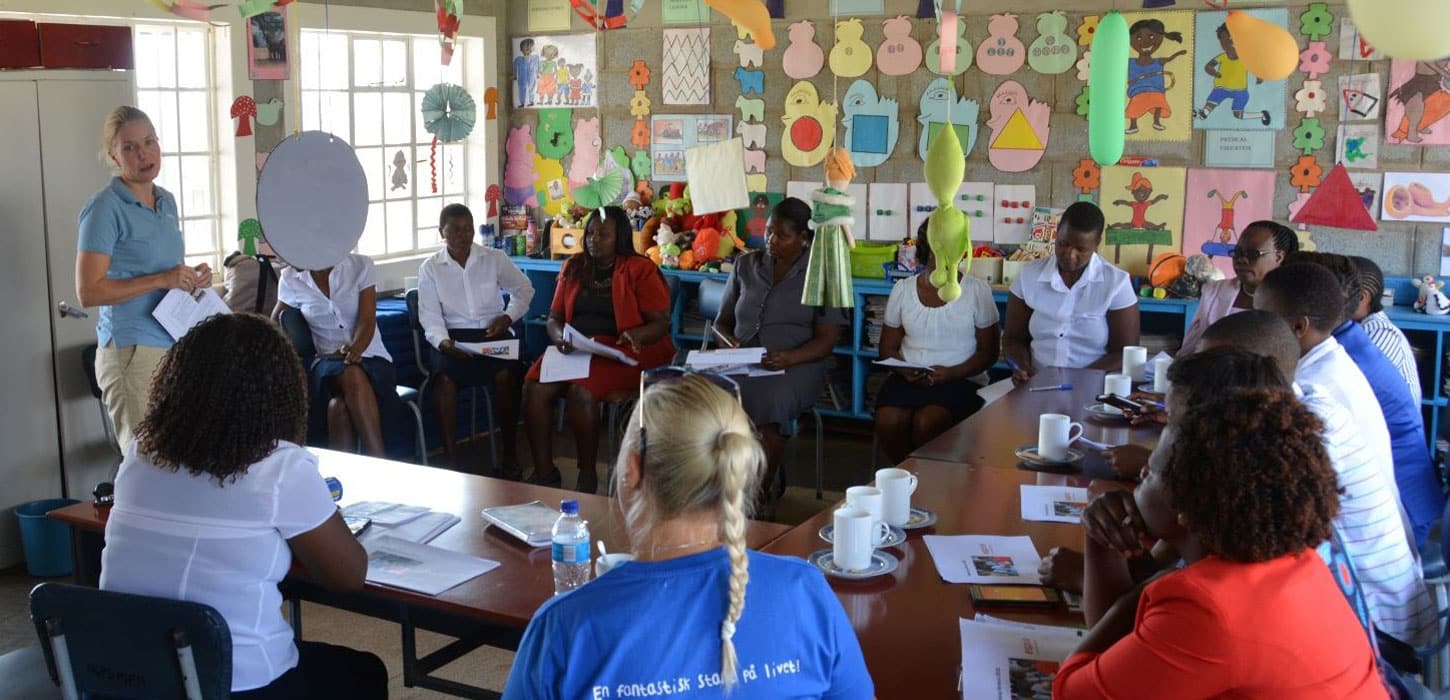 Pedagoger fra barnehagen i Zimbabwe og Espira sitte i et klasserom og har en workshop. På en av veggene er det hengt opp fargerike ark og plakater og i taket henger det ballonger. Foto: Espira