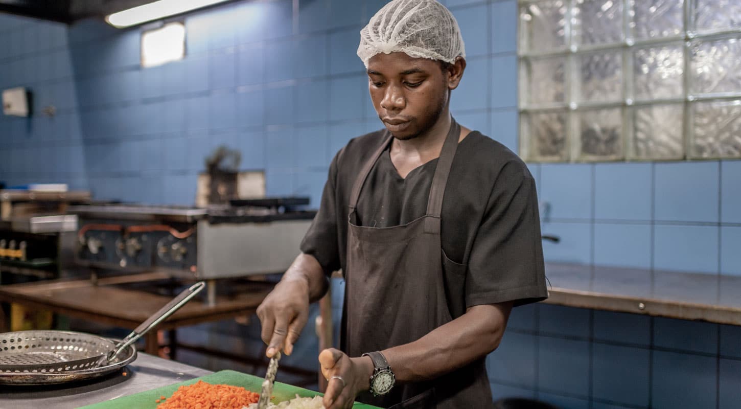 Marcos fra Malawi står i et kjøkken med blå fliser på veggen og skjærer gulrøtter. Han har svart skjorte uten krage og grått forkle, på hodet har han et nett, Foto: Cornel van Heerden