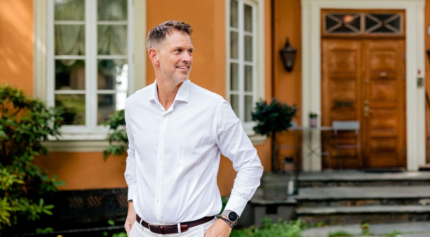 Øyvind Gabrielsen, franchisetaker og eier for Aktiv Eiendom Oslo, har kort hår med gråstenk i tinningen, hvit skjorte og lys bukse. Han står foran et gult hus med hvite karmer og brun dør. Foto Ronja Sagbakken