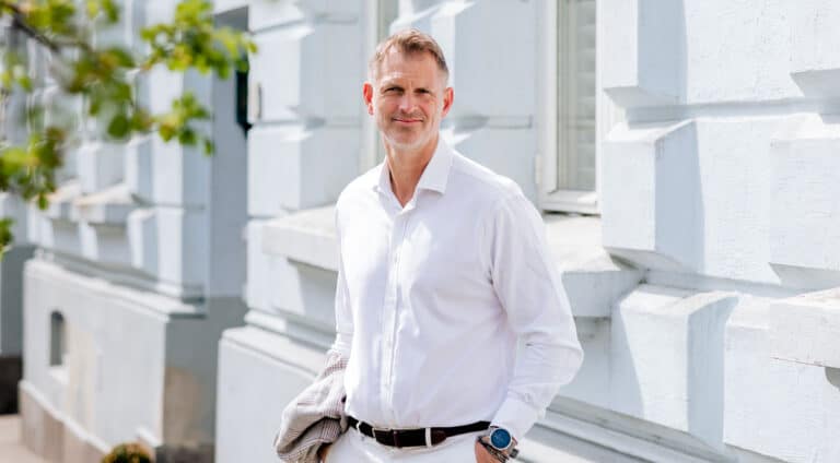 Øyvind Gabrielsen, franchisetaker og eier for Aktiv Eiendom Oslo, har kort hår med gråstenk i tinningen, hvit skjorte og lys bukse. Han står foran et bygg med hvite murvegger. Foto Ronja Sagbakken