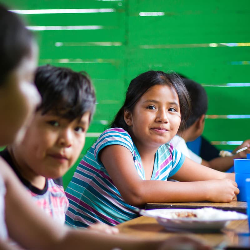 Flere barn, med mørkt hår og t-skjorte, sitter ved et bord og spiser. Det er en grønn vegg i bakgrunnen og de har tallerkener blå kopper foran seg. Foto: Danielle Pereria