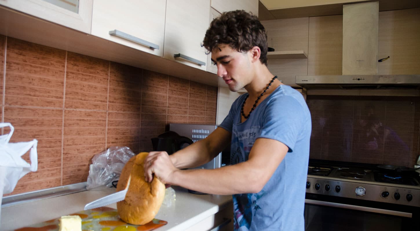 Ung gutt med mørkt, kort, bølget hår og blå t-skjorte står ved kjøkkenbenken og skjærer brød. Foto: Katerina Ilievska