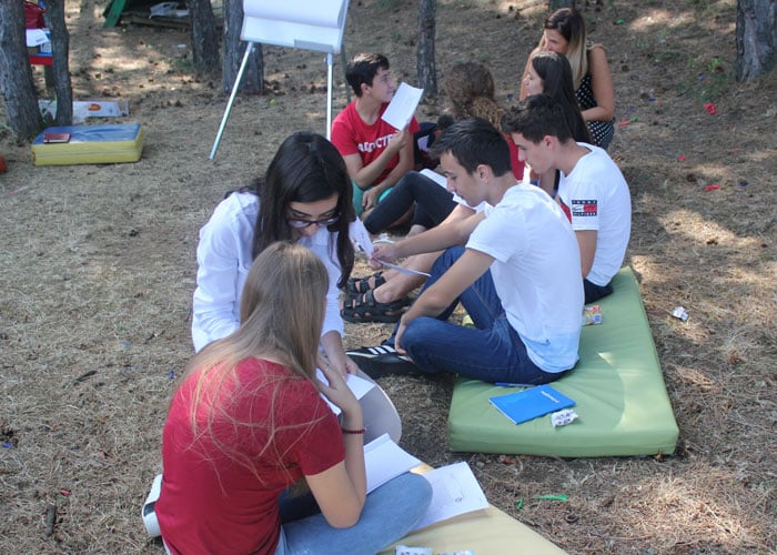 En gruppe ungdom sitter på bakken på madrasser og jobber med oppgaver i fellesskap. Veilederen er sitter på huk og forklarer. Foto: SOS-barnebyer