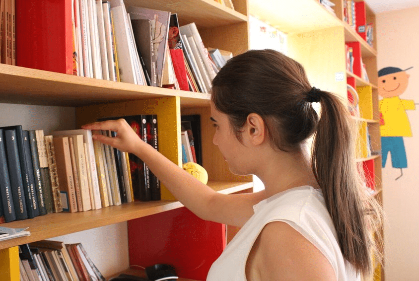 Ei tenåringsjente står foran ei bokhylle og ser på bøkene. Hun har langt mørkt hår i hestehale og hvit t-skjorte uten ermer. Illustrasjonsfoto: Katerina Ilievska