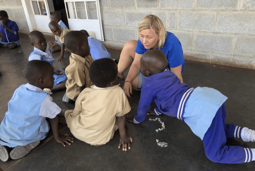 Pedagog Tilla Minge, med lyst halvlangt hår, blå t-skjorte og shorts, sitter på gulvet og leker med flere barn fra barnehagen i Zimbabwe. Foto: Espira