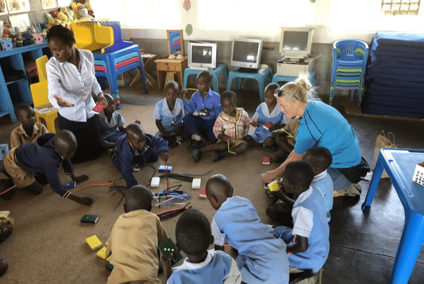 Pedagog Veronica Egra fra Espira, med lyst langt hår i hestehale, solbriller på hode, blå t-skjorte og jeans sitter på huk inne i barnehagen og leker med barna i barnehagen i Zimbabwe. Foto: Espira