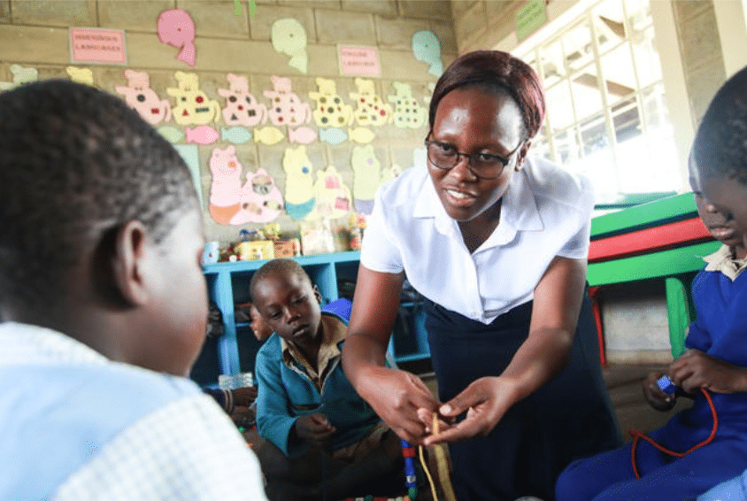 Pedagog Pauline Mhako fra SOS-barnehagen i Zimbabwe, med rødlig glatt hår i en topp, hvit skjorte og mørkeblått skjørt viser noen barn en leke. Foto: Espira