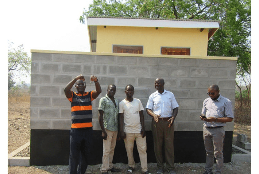 Fem menn står foran murveggen som skjuler det nye toalettanlegget. Foto SOS-barnebyer 