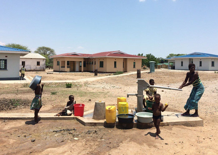 En kvinne fyller vannkanner med vannpumbpen, to barn står sammen med henne, i bakgrunnen er husene i SOS-barnebyer Ngabu. Foto: SOS-barnebyer