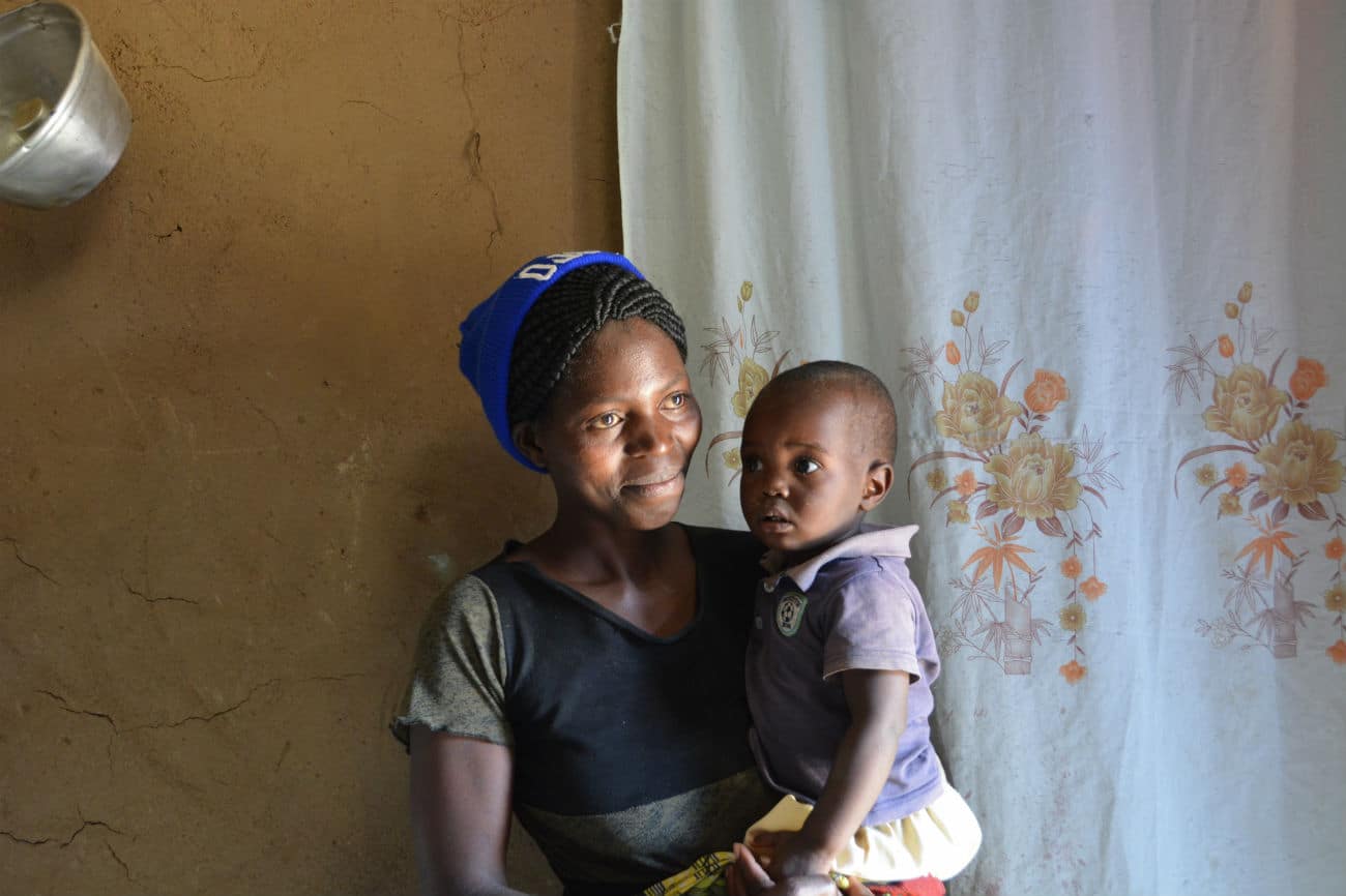 En mor, som er en del av det forebyggende arbeidet i Huambo, står i et rom med gule murvegger, og blomstrete gule og hvite gardiner, hun har fletter satt opp i en topp, blå lue, mørk t-skjorte. Den lille sønnen hennes sitter på armen, han har lilla kortermet skjorte. Foto: Turid Weisser