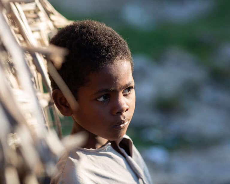 Portrettbilde av gutt fra Tanzania med mørkt, kort hår og lysegrå genser med knapper, står og lener seg inntil en stråvegg. Illustrasjonsfoto: Lars Just